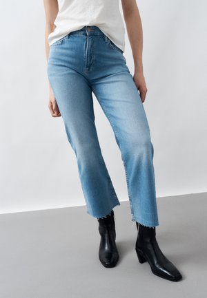 Henkilö, jolla on vaaleansiniset polvipituiset farkut, joissa on rispaantuneet reunat, valkoinen sisäänsolmittu paita ja mustat korolliset nilkkurit, seisoo harmaalla lattialla.