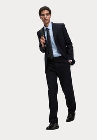 Męski ciemnoniebieski garnitur o dopasowanym kroju, biała koszula i krawat w kropki. W komplecie czarna torba na ramię i błyszczące czarne buty.