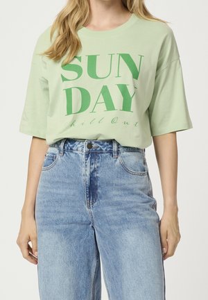 Kvinde iført en lys grøn oversized T-shirt med teksten "SUNDAY chill out" i grønt, stukket ned i højttaljede lyseblå jeans.