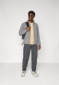 Grijze zip-up jack, beige T-shirt en donkergrijze broek. Kenmerken zijn onder andere een gestructureerde stof, een logo op het jack en grijze sportschoenen.