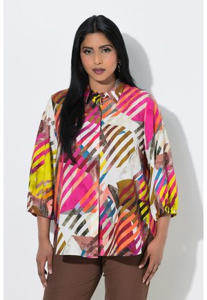 Femme aux longs cheveux noirs portant un chemisier coloré à motif géométrique avec des manches bouffantes et un pantalon marron, debout sur un fond blanc.