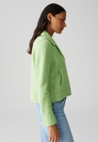 Giacca verde strutturata con colletto e due tasche laterali. Presenta un taglio corto e maniche lunghe, indossata sopra jeans azzurri chiari.