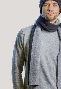 Sciarpa a maglia grigia scura drappeggiata su un maglione grigio chiaro. La sciarpa presenta una texture a coste e un accento blu a contrasto. Materiale accogliente e vestibilità aderente.