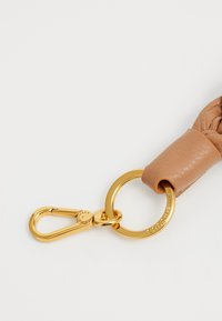 Bracelet en cuir marron avec une texture tissée, présentant des éléments en métal doré, un fermoir et un anneau circulaire gravé du nom de la marque.