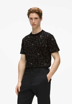 Jeune homme aux cheveux châtain clair portant une chemise noire à manches courtes à motifs et un pantalon noir, debout avec une main dans sa poche.