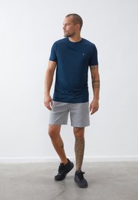 Camisola desportiva de manga curta azul-marinho com decote redondo, combinada com calções cinza. O modelo usa ténis pretos e exibe uma tatuagem geométrica.