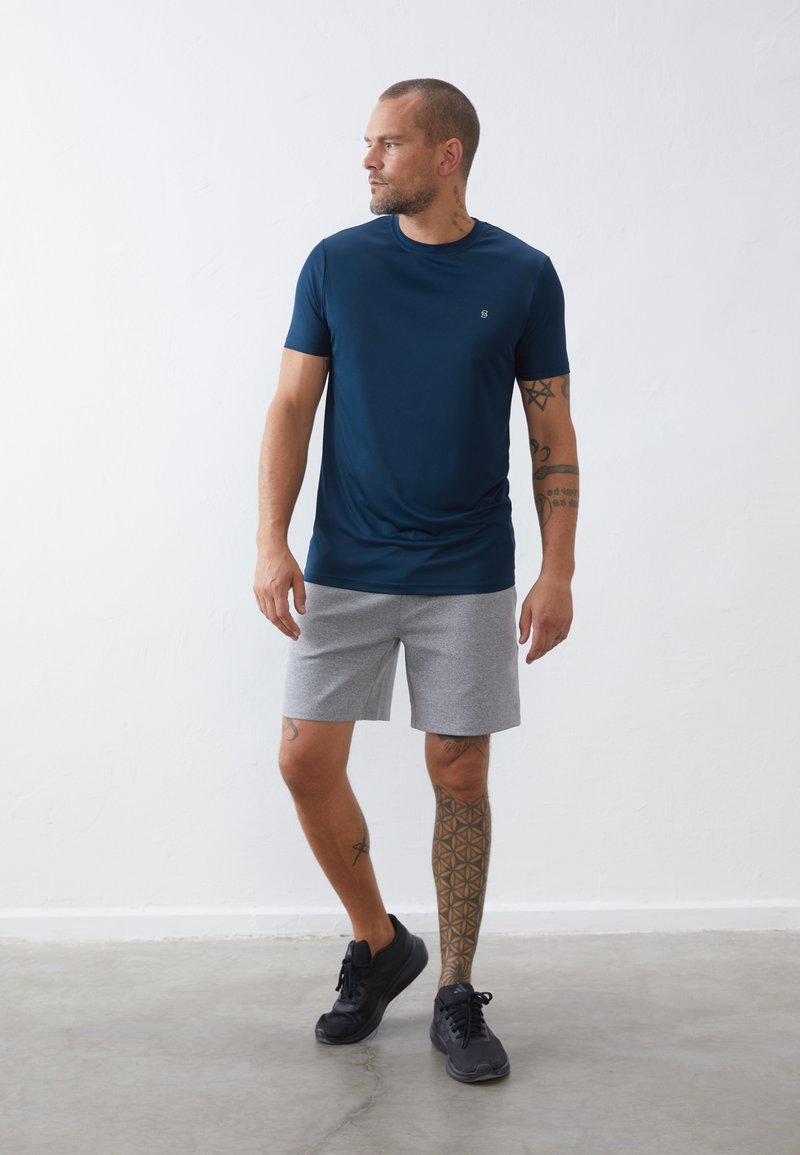 Camiseta atlética de manga corta color azul marino con cuello redondo, combinada con pantalones cortos grises. El modelo lleva zapatillas negras y muestra un tatuaje geométrico.