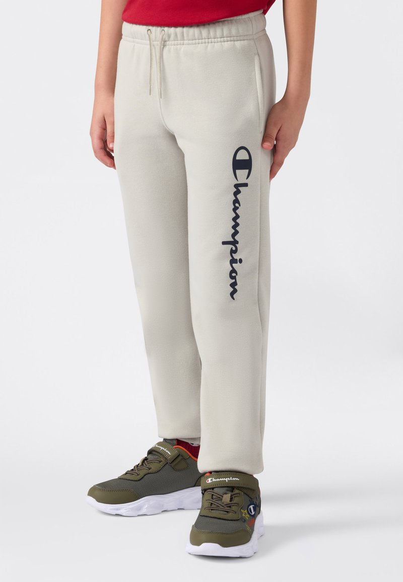 Barn som har på seg beige sweatshirtbukser med svart "Champion"-logo på benet og olivengrønne Champion-sko med hvite såler.