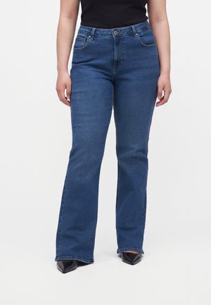 Partie inférieure du corps d'une personne portant un jean bootcut bleu, des chaussures noires pointues et un haut noir sans manches sur un fond uni.