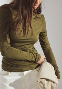 Olivu zaļš garām piedurknēm tops ar savilktu dekoltē, kombinēts ar baltām džinsām un turēts beiges somas. Gluda, pieguļoša auduma tekstūra.