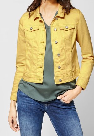 Kobieta w żółtej dżinsowej kurtce na guziki, na zielonym topie z dekoltem w serek i niebieskich dżinsach, z jedną ręką w kieszeni.