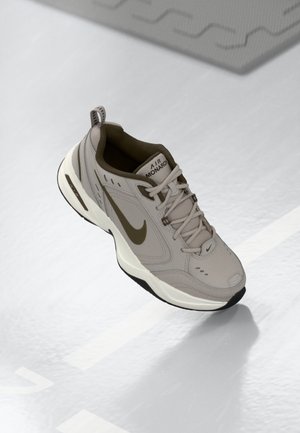 Hellgrauer Nike Air Monarch Sneaker mit olivgrünem Swoosh und Akzenten, weißer Sohle und Schnürsenkeln, gezeigt vor einem reflektierenden hellen Hintergrund.