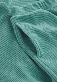 Tessuto a coste in un morbido color teal, con una superficie testurizzata e una leggera elasticità. Il materiale cade naturalmente con cuciture e pieghe visibili.