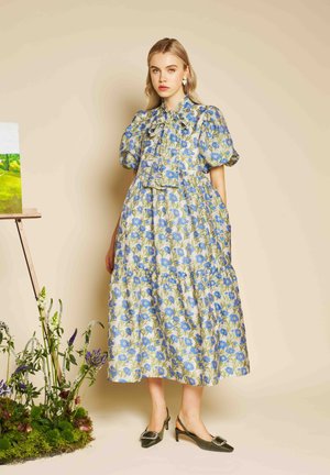 Blonde woman wearing a blue floral puff-sleeve midi dress and black heeled slingback shoes, standing near flowers and a painting.