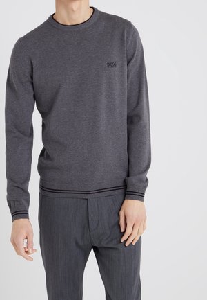 Mand iført en tætsiddende mørkegrå langærmet sweater med BOSS-logo og matchende mørkegrå bukser mod en ensfarvet baggrund.
