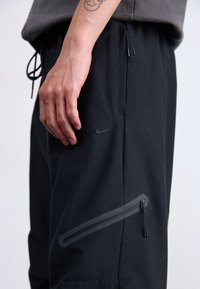 Detailný záber na ruku odpočíva vedľa čiernych nohavíc Nike s diagonálnou zipsovou kapsou a viditeľným šnúrkovým pásom.