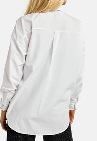 Valge nööpidega pluus, millel on pikad varrukad, tagumisel osal plisseeritud detail ja kõverdatud serv. Kangas tundub sile ja veidi läikiva viimistlusega.