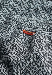 Zwart-witte shorts met herhaalde "SUPERDRY" branding. Elastische tailleband met een oranje label. Glad materiaal met een bedrukt patroon.