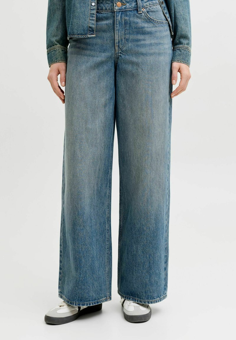 Persoană purtând blugi largi din denim albastru și adidași albi, stând în fața unui fundal simplu, deschis la culoare.