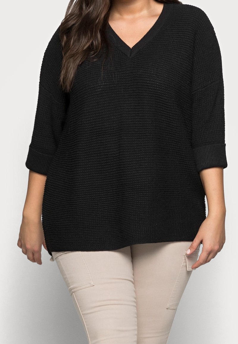 Pull noir à maille lâche avec col en V, manches 3/4 et poignets côtelés. Tissu texturé ; associé à un pantalon beige clair.