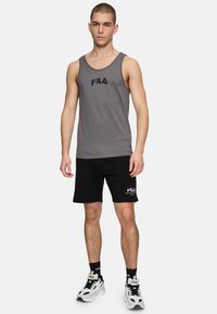 Camiseta sin mangas gris con un logotipo negro, combinada con pantalones cortos negros que tienen un detalle de logotipo. Lleva zapatillas en blanco y negro.