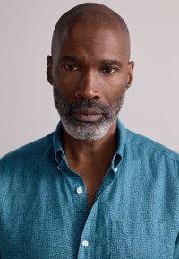 Camisa turquesa abotonada con patrones geométricos, que presenta un cuello y dos botones blancos en la parte delantera. Textura suave y diseño ajustado.