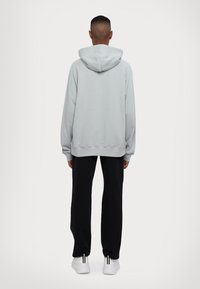 Άτομο που στέκεται με το πρόσωπο στραμμένο μακριά, φορώντας μια ανοιχτή γκρι φούτερ, μαύρα παντελόνια και λευκά αθλητικά παπούτσια, μπροστά σε έναν καθαρό λευκό φόντο.