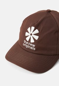 Boné castanho feito de tecido com uma aba plana, logotipo de flor branca bordado e texto a dizer "os novos originais" na parte da frente.