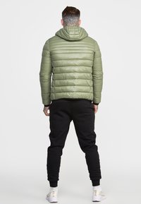 Man with neck tattoo wearing a green puffer jacket, black joggers, white socks, and white sneakers standing facing away on a white background.
