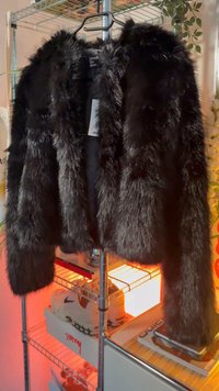 Crna jakna od umjetne krzno s dugim rukavima, bez ovratnika i teksturiranim završetkom. Obješena na crnom vješalu protiv osvijetljenog pozadine.