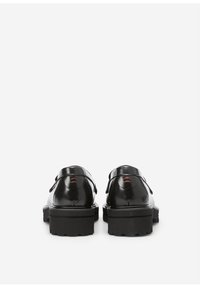 Un par de mocasines de cuero negro con suelas de goma gruesas, vistos desde atrás contra un fondo blanco liso.