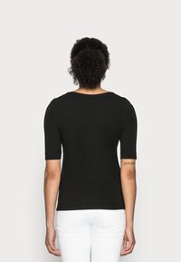 Personne aux cheveux bouclés portant un haut noir à manches courtes et un pantalon blanc, vue de dos sur un fond uni.