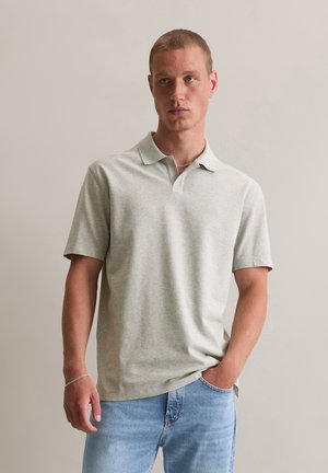Lichtgrijs polo shirt van zacht materiaal, met een klassieke kraag en korte mouwen. G gedragen met blauwe jeans, handen in de zakken.