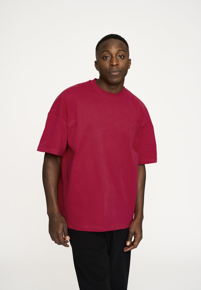 Rød oversized bomulds t-shirt med rund hals og korte ærmer, parret med sorte bukser, på en neutral baggrund.