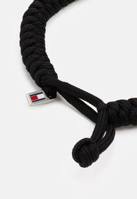 Svart flettet tauarmbånd med en sølvfarget hardware-detalj som har et rektangulært logo-merke i rødt og blått. Endene er knyttet i knuter.