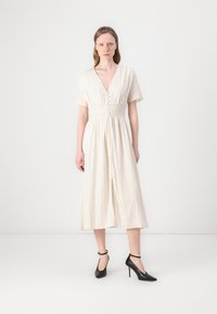 Abito color crema con maniche corte, scollatura a V e chiusura frontale con bottoni. Il tessuto ha una sottile texture verticale. Abbinato a scarpe con tacco alto nere.