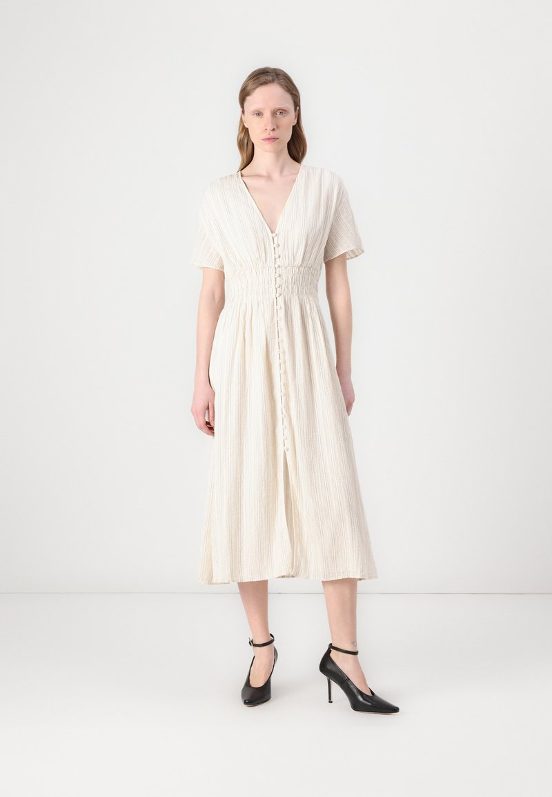 Abito color crema con maniche corte, scollatura a V e chiusura frontale con bottoni. Il tessuto ha una sottile texture verticale. Abbinato a scarpe con tacco alto nere.