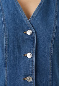 Vestido de ganga azul com decote em V, apresentando cinco botões de metal prateado e costuras visíveis. Design estruturado com uma textura suave.