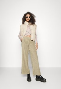 Colete puffer bege por cima de uma blusa de mangas longas listrada em várias cores, combinado com umas calças de veludo cotelê verdes largas e texturizadas e botas de plataforma pretas.