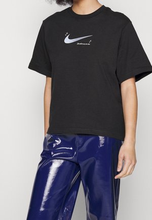 Osoba nosząca czarną koszulkę z srebrnym logo Nike i napisem, w połączeniu z błyszczącymi ciemnoniebieskimi spodniami.