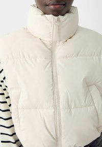 Beige puffer vest with a high collar, front zipper, and two side pockets. The fabric appears smooth, with subtle stitching detailing.