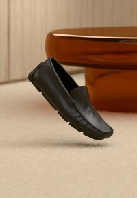 Μαύρο μοκασίνι από δέρμα που αιωρείται πάνω από μπεζ χαλί δίπλα σε στρογγυλό τραπεζάκι από γυαλί χρώματος κεχριμπαριού.