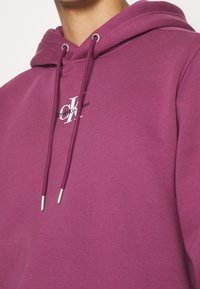 Gros plan sur une personne portant un sweat à capuche violet avec des cordons et un petit logo blanc "CK" sur la poitrine.