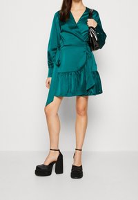 Tyrkysové saténové zavinovací šaty s dlouhými rukávy, volánkovým lemem a detailem zavazování v oblasti pasu; doplněné černými platformovými lodičkami.