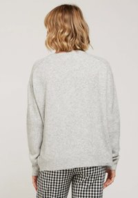 Pull léger gris clair avec une texture douce, des poignets et un ourlet côtelés. Présente une coupe décontractée et des manches raglan. Pantalons à carreaux noir et blanc visibles en dessous.