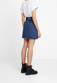 Denim-Minirock mit hoher Taille, blauer Farbe und zwei Gesäßtaschen, kombiniert mit einem weißen Hemd mit hochgekrempelten Ärmeln und schwarzen Schnürstiefeln.