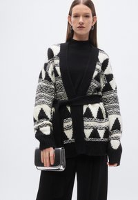 Πλεκτή ασπρόμαυρη ζακέτα με γεωμετρικά σχέδια, ανοιχτή μπροστά, με μακριά μανίκια και ζώνη στο ίδιο σχέδιο, κρατώντας μια γκρι τσάντα φάκελο.