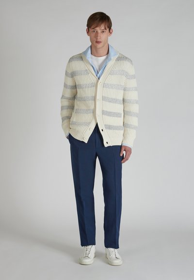 Sweter w paski w kolorze kremowym i szarym z kołnierzem szalowym, z zapięciem na guziki, zestawiony z granatowymi spodniami i białymi sneakersami. Gładka tekstura.