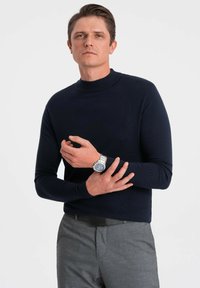 Marineblauer Langarmpullover mit hohem Kragen, aus einem gerippten Stoff. Kombiniert mit grauen Hosen und einer silbernen Armbanduhr.