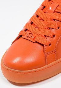 Světlé oranžové tenisky s hladkým koženým svrškem, plochými šňůrkami a kulatou špičkou. Mají logo "MK" na jazyku a oranžovou gumovou podrážku.